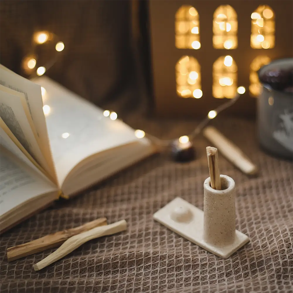 Image apaisante et un peu mystique. On peut voir en premier plan un bâton de Palo Santo sur un support. D'autres bâtons de Palo Santo sont posés sur un tissus, un livre est ouvert. L'arrière plan est flouté et on peut voir une guirlande lumineuse et de nombreuses petites lumières jaunes.