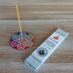 Image de l'encens Ayurvedic sauge blanche en bâtonnets. Cette boite d'encens est posée à côté d'un porte encens rond coloré sur lequel il y a un bâtonnet d'encens à la sauge blanche. L'image est prise sur un fond zen. Ces produits sont en vente chez Zen Aatma - boutique zen et ésotérique.