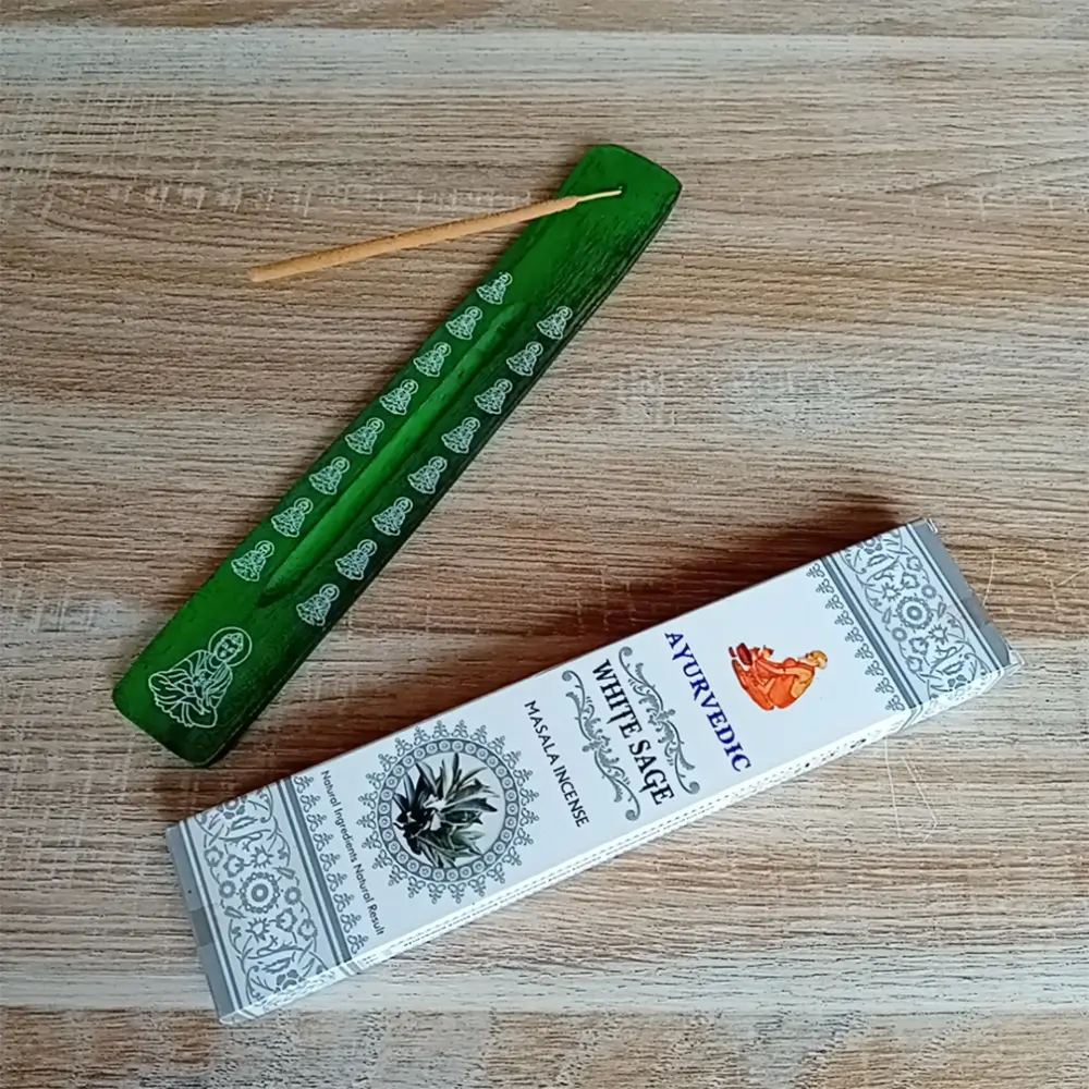 Image de l'encens Ayurvedic sauge blanche en bâtonnets. Cette boite d'encens est posée à côté d'un porte encens en bois coloré en vert foncé sur lequel il y a un bâtonnet d'encens. L'image est prise sur un fond zen. Ces produits sont en vente chez Zen Aatma - boutique zen et ésotérique.