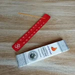 Image de l'encens Ayurvedic sauge blanche en bâtonnets. Cette boite d'encens est posée à côté d'un porte encens en bois coloré en rouge sur lequel il y a un bâtonnet d'encens. L'image est prise sur un fond zen. Ces produits sont en vente chez Zen Aatma - boutique zen et ésotérique.