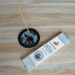 Image de l'encens Ayurvedic sauge blanche en bâtonnets. Cette boite d'encens est posée à côté d'un porte encens rond noir avec des motifs zen. Sur ce porte encens, il y a un bâtonnet d'encens. L'image est prise sur un fond zen. Ces produits sont en vente chez Zen Aatma - boutique zen et ésotérique.