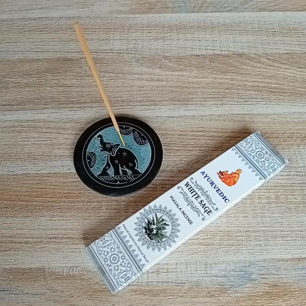 Image de l'encens Ayurvedic sauge blanche en bâtonnets. Cette boite d'encens est posée à côté d'un porte encens rond noir avec des motifs zen. Sur ce porte encens, il y a un bâtonnet d'encens. L'image est prise sur un fond zen. Ces produits sont en vente chez Zen Aatma - boutique zen et ésotérique.