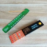 Image de l'encens Tribal Soul Copal blanc en bâtonnets. Cette boite d'encens est posée à côté d'un porte encens artisanal en bois vert foncé sur lequel il y a un bâtonnet d'encens au Copal blanc. L'image est prise sur un fond zen. Ces produits sont en vente chez Zen Aatma - boutique zen et ésotérique.