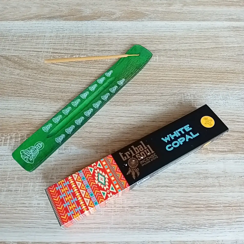 Image de l'encens Tribal Soul Copal blanc en bâtonnets. Cette boite d'encens est posée à côté d'un porte encens artisanal en bois vert foncé sur lequel il y a un bâtonnet d'encens au Copal blanc. L'image est prise sur un fond zen. Ces produits sont en vente chez Zen Aatma - boutique zen et ésotérique.
