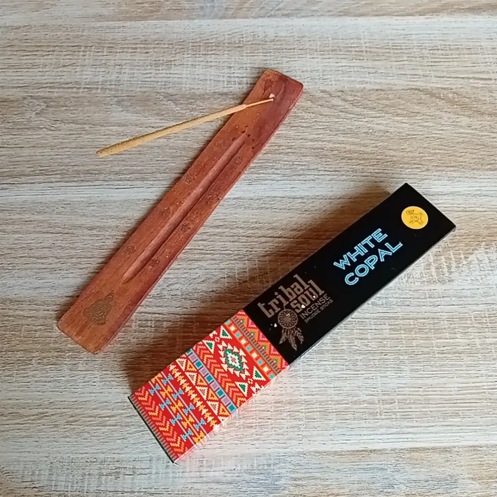 Image de l'encens Tribal Soul Copal blanc en bâtonnets. Cette boite d'encens est posée à côté d'un porte encens artisanal en bois sur lequel il y a un bâtonnet d'encens au Copal blanc. L'image est prise sur un fond zen. Ces produits sont en vente chez Zen Aatma - boutique zen et ésotérique.