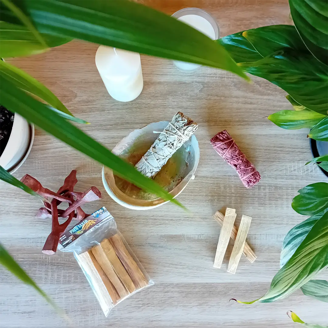 La photo est prise de près sur un fond en bois clair. On peut voir une coquille d'ormeau posé sur un trépied Cobra naturel. Dans la coquille se trouve un fagot de sauge blanche. A côté on voit un fagot de sauge sang de dragon, des bâtons de Palo Santo, un trépied cobra laqué. Ces produits font partie du pack rituel de purification naturelle de la boutique Zen Aatma. L'ambiance de l'image est très zen. On peut trouver ces produits dans la boutique en ligne d'articles zen et ésotériques Zen Aatma.