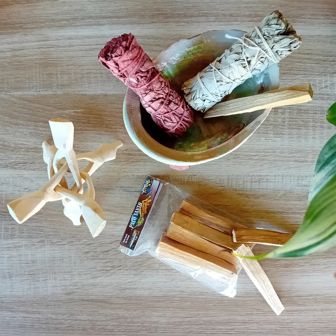 La photo est prise de près sur un fond en bois clair. On peut voir une coquille d'ormeau posé sur un trépied Cobra laqué. Dans la coquille se trouve un fagot de sauge blanche, un fagot de sauge sang de dragon et un bâton de Palo Santo. A côté on voit des bâtons de Palo Santo, un trépied cobra naturel. Ces produits font partie du pack rituel de purification naturelle, un des nombreux packs de la boutique Zen Aatma. L'ambiance de l'image est très zen. On peut trouver ces produits dans la boutique en ligne d'articles zen et ésotériques Zen Aatma.