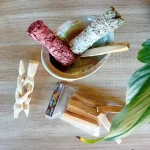 La photo est prise de près sur un fond en bois clair. On peut voir une coquille d'ormeau posé sur un trépied Cobra laqué. Dans la coquille se trouve un fagot de sauge blanche, un fagot de sauge sang de dragon et un bâton de Palo Santo. A côté on voit des bâtons de Palo Santo, un trépied cobra naturel. Ces produits font partie du pack rituel de purification naturelle, un des nombreux packs de la boutique Zen Aatma. L'ambiance de l'image est très zen. On peut trouver ces produits dans la boutique en ligne d'articles zen et ésotériques Zen Aatma.