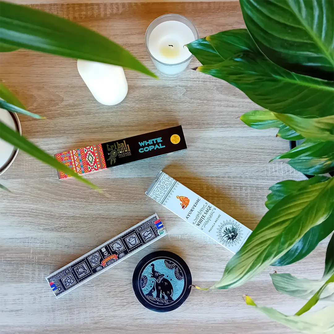 On peut voir une boite d'encens Tribal Soul Copal blanc, une boite d'encens Ayurvedic sauge blanche, une boite d'encens Satya super hit et un porte encens noir en pierre. Ces produits font partie du pack voyage spirituel de la boutique Zen Aatma. Il y a aussi deux bougies et des plantes vertes. L'ambiance de l'image est très zen. On peut trouver ces produits dans la boutique en ligne d'articles zen et ésotériques Zen Aatma.